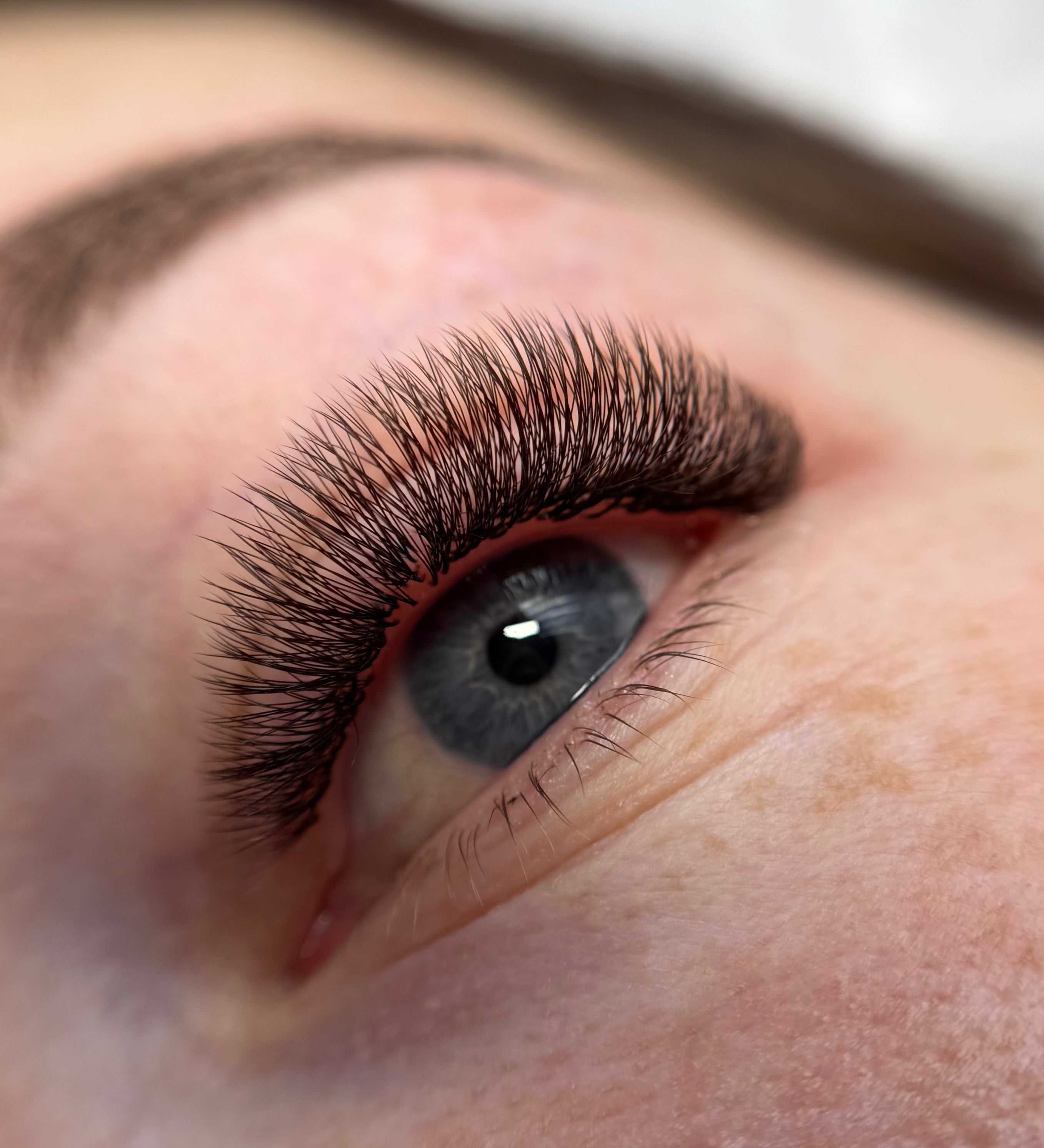 Close-up of a person's eye with long, dark brown, and well-defined eyelash extensions applied, showing the lashes in sharp detail against natural skin.