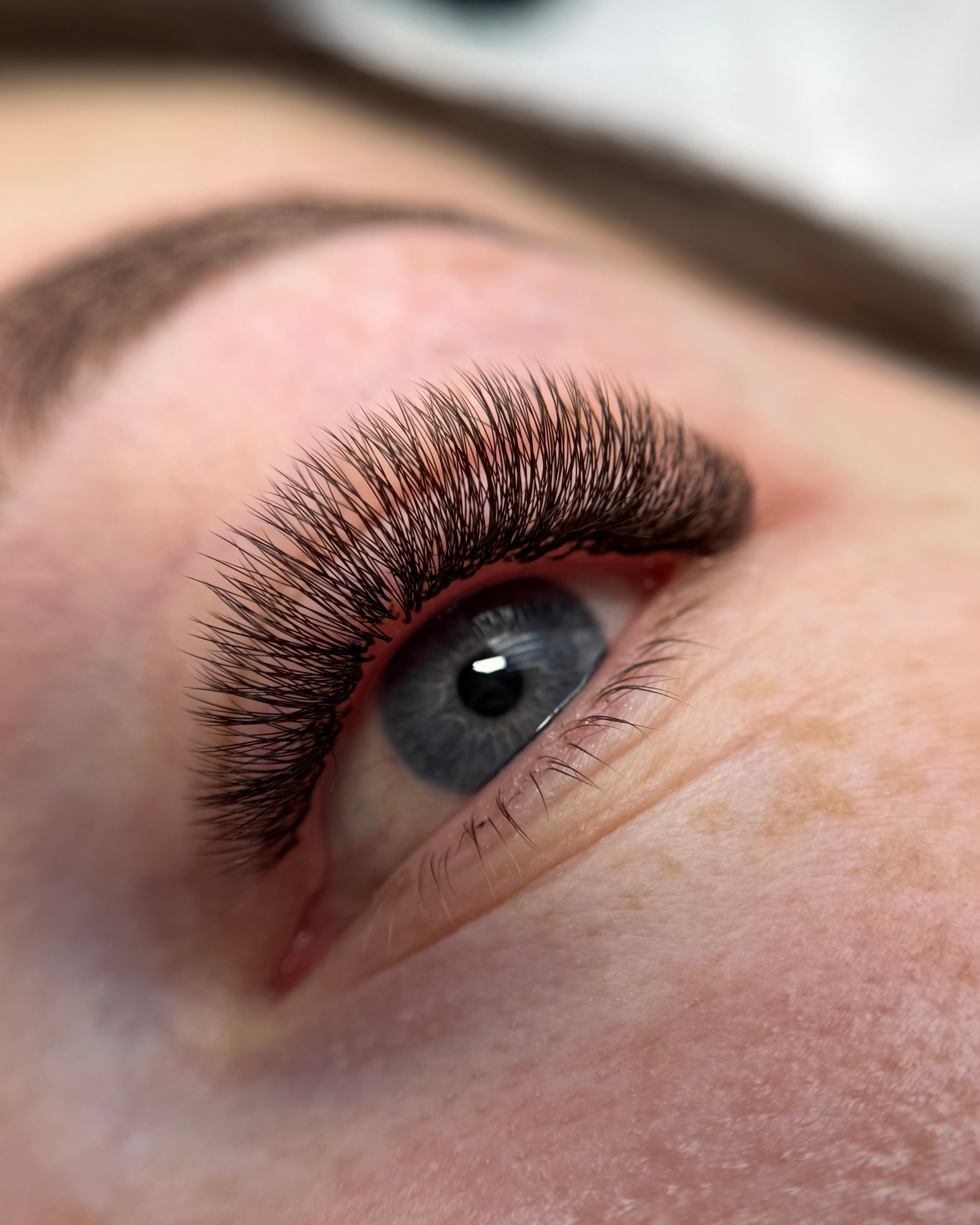 Close-up of a person's eye with long, dark brown, and well-defined eyelash extensions applied, showing the lashes in sharp detail against natural skin.