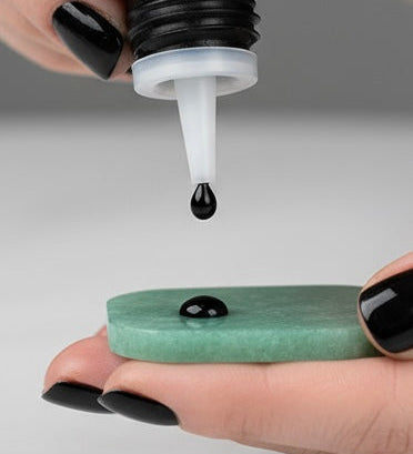 A close-up of a hand with black manicured nails holding a small green jade stone while another hand drops a black lash glue drop onto the stone.