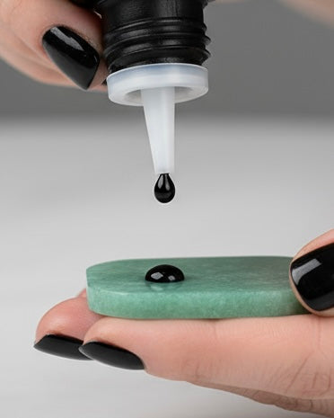 A close-up of a hand with black manicured nails holding a small green jade stone while another hand drops a black lash glue drop onto the stone.